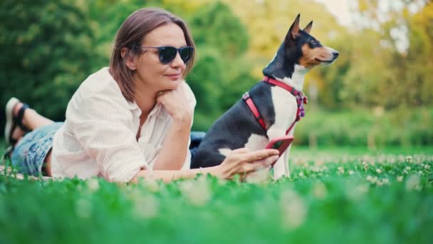 Mladá dospělá žena leží v parku na kostce se svým psem Basendžim — Stock video