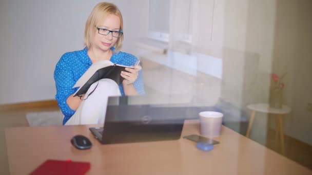 Volwassen blonde vrouw het maken van notities in haar notebook tijdens het kijken naar een webinar — Stockvideo