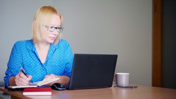 Erwachsene blonde Frau macht sich Notizen in ihrem Notizbuch, während sie ein Webinar anschaut — Stockvideo