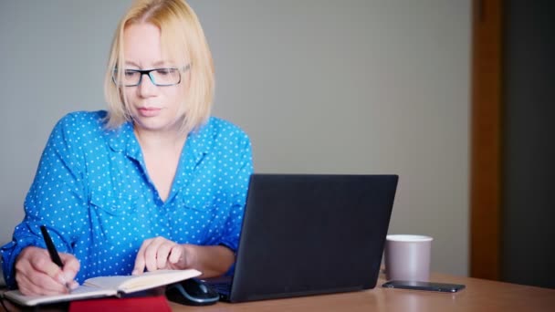 Volwassen blonde vrouw het maken van notities in haar notebook tijdens het kijken naar een webinar — Stockvideo