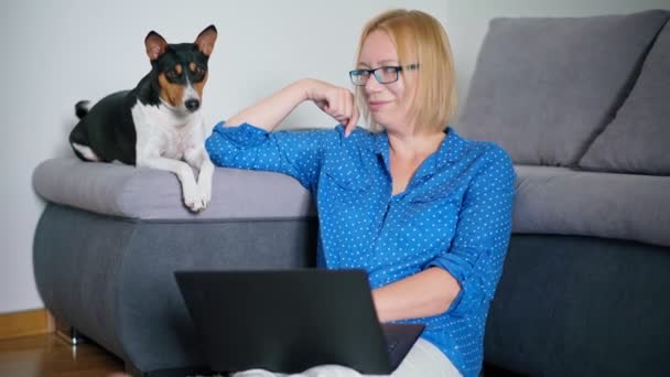 Une femme travaillant sur un ordinateur portable tout en étant assise sur le sol avec son chien mignon — Video