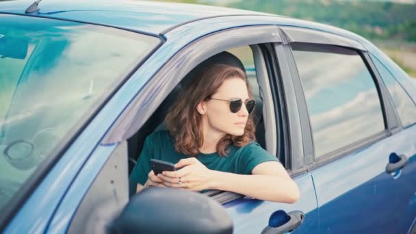 Genç bir kadın arabada otururken telefonunu kullanıyor ve manzaranın tadını çıkarıyor. — Stok video