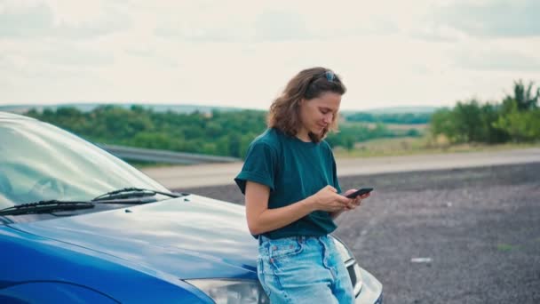 Молодая женщина использует свой телефон опираясь на бампер автомобиля и наслаждаясь видом — стоковое видео