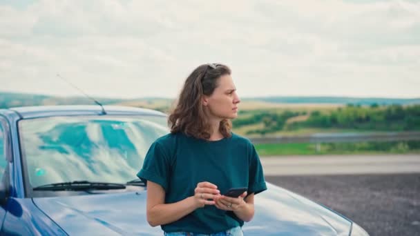 Mujer joven utiliza su teléfono apoyado en el parachoques de un coche y disfrutar de la vista — Vídeos de Stock