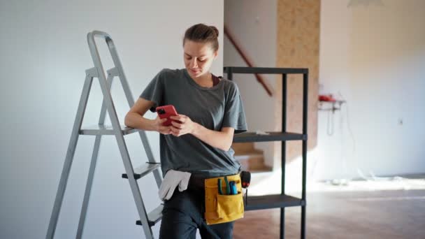 Eine professionelle Reparaturfachfrau benutzt ihr Telefon, während sie sich auf eine Leiter stützt — Stockvideo