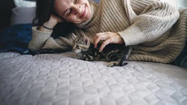 Genç bir kadın, yatakta uzanırken küçük sevimli bir kediyi okşar.