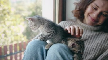 Genç bir kadın, bir kır evinin pencere eşiğinde oturan sevimli kedilerle oynuyor.