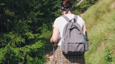 Güneşli bir yaz gününde sırt çantalı ve sırıkla vahşi bir patikada yürüyen genç bayan yürüyüşçünün arka görüntüsü. Macera, özgürlük ve aktif seyahat yaşam tarzı konsepti.