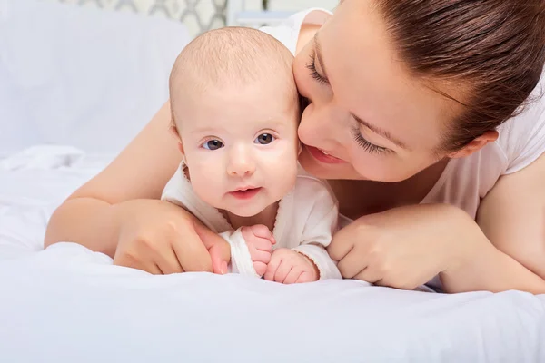 Happy baby girl lying near her mother on a white bed. Girls looking at each other. — Stock Photo, Image