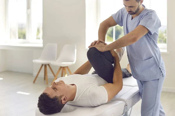 Poważny lekarz pomaga męskiemu sportowcowi wykonywać ćwiczenia fizyczne podczas rehabilitacji po urazie nogi — Zdjęcie stockowe
