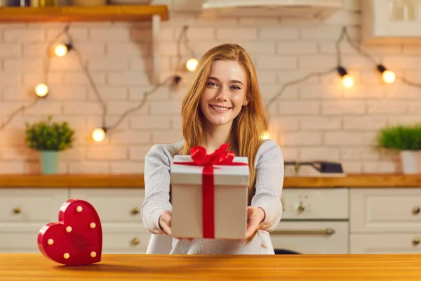 Smiling young woman holding out Valentine present sitting at table in the kitchen — Stock Photo, Image