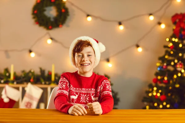 Щасливий хлопчик у традиційному різдвяному джемпері та шапці Санта сидить за столом та посміхається — стокове фото