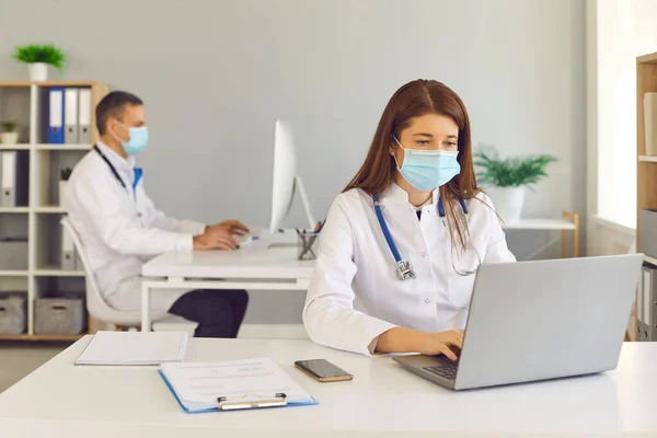 Dokter bertopeng wajah medis yang bekerja pada komputer yang duduk di meja di kantor — Stok Foto