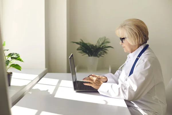 Starsza uśmiechnięta kobieta lekarz siedzi z laptopem i komunikuje się z pacjentem online — Zdjęcie stockowe