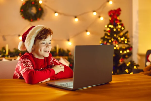 Szczęśliwy chłopiec w świątecznym czerwony kapelusz Mikołaja i sweter siedzi i ogląda Boże Narodzenie film online na laptopie — Zdjęcie stockowe
