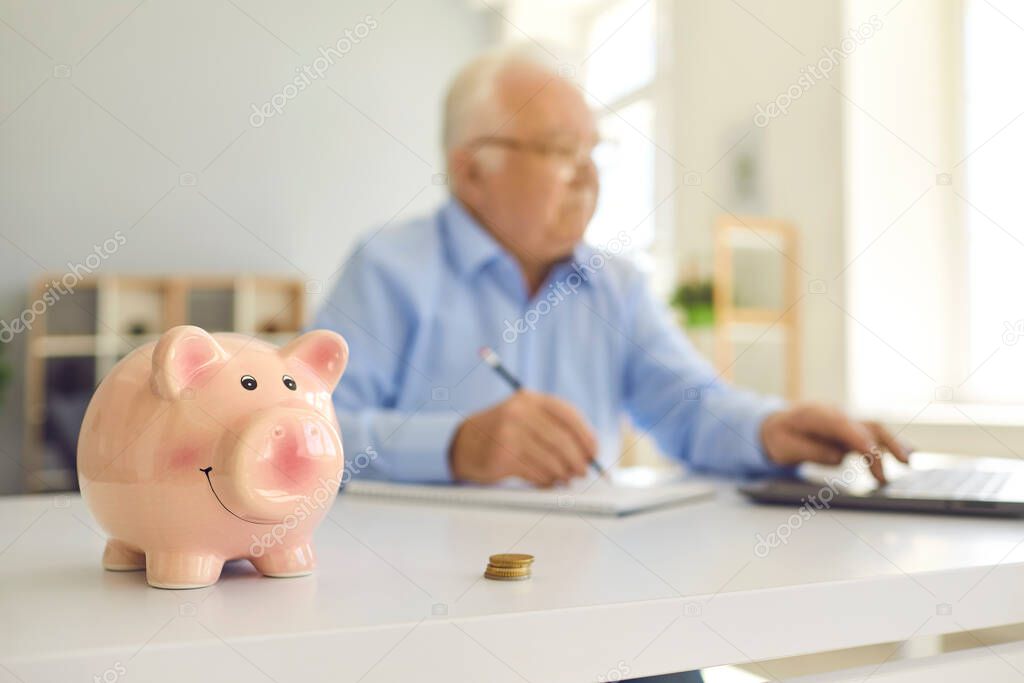 Piggy bank standing on desk with blurred retired man using laptop and doing accounting in background