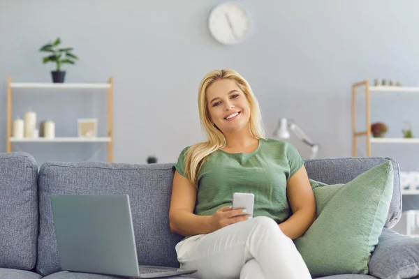Happy woman sitting on couch with laptop, holding mobile phone in hand and looking at camera — Stock Photo, Image
