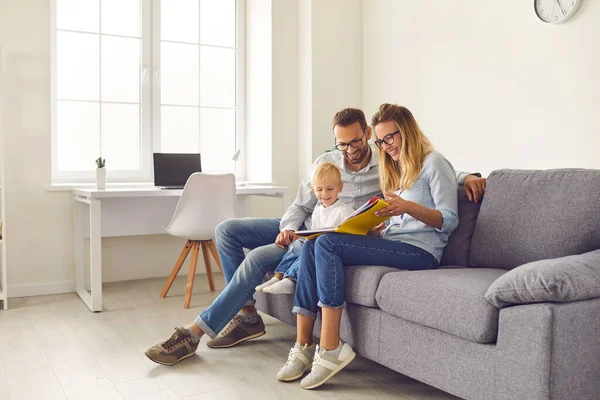 Gelukkige ouders en kind zitten op de bank, lezen boek van verhalen of kijken door familie fotoalbum — Stockfoto