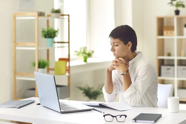 Mulher pensiva sentada na mesa com laptop e notebook assistindo vídeo educativo ou leitura ebook — Fotografia de Stock