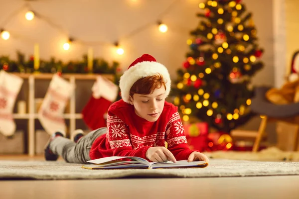 Concentrated boy in red Christmas clothing and hat lying on floor and reading book — Stock Photo, Image