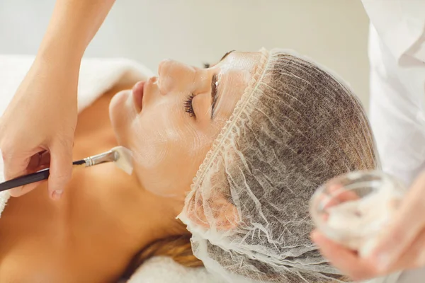 Manos de esteticista aplicando mascarilla hidratante con cepillo cosmético a mujer joven relajada —  Fotos de Stock