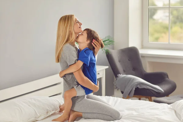 Glückliche Mutter sitzt im Schlafzimmer auf dem Bett und umarmt ihren kleinen Sohn — Stockfoto