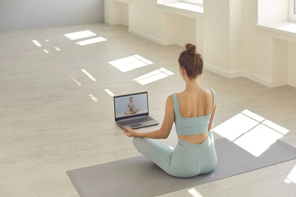 Woman sitting legs crossed on floor at home watching yoga video lesson on laptop