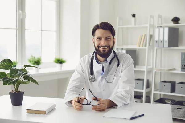 Szczęśliwy brodaty lekarz siedzi przy stole w gabinecie medycznym. — Zdjęcie stockowe