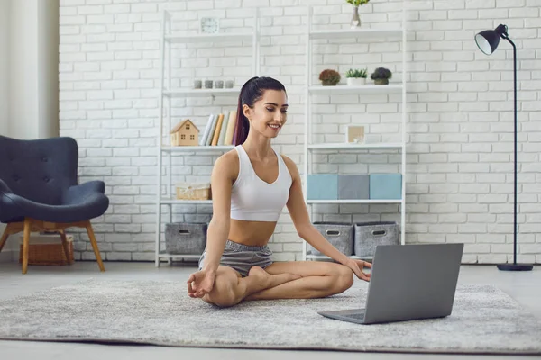 Girl sitting on carpet in lotus position and practicing yoga during online lesson on laptop at home