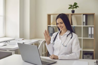 Hastane ofisindeki dost canlısı doktor bilgisayar ekranına el sallıyor ve video görüşmesi yapıyor.
