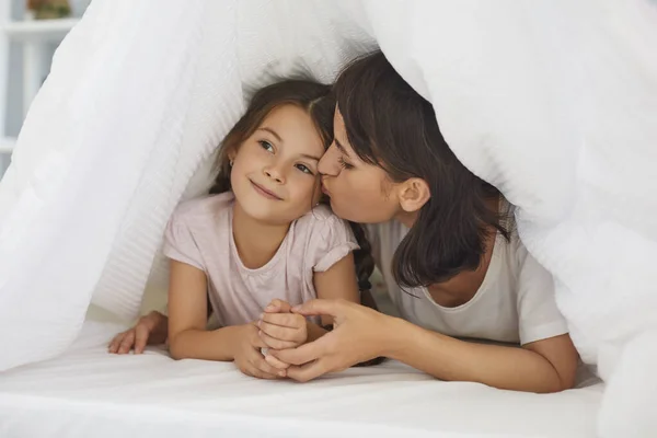 Liebevolle junge Mutter küsst ihre kleine Tochter auf dem Bett unter der Decke im Schlafzimmer. — Stockfoto