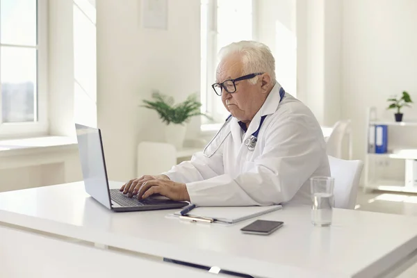 Poważny lekarz medycyny ogólnej z siwymi włosami pracujący na laptopie siedzącym przy biurku w biurze szpitalnym — Zdjęcie stockowe