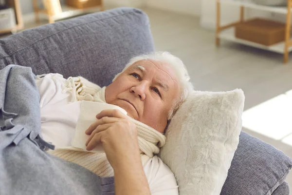 Malade homme âgé avec une écharpe enroulée autour de son cou se trouve sur un canapé à la maison et est traitée. — Photo