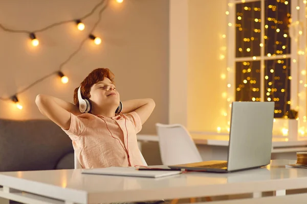 Tranquilo chico con los ojos cerrados escucha música en los auriculares delante de la computadora portátil recostado en la silla. — Foto de Stock