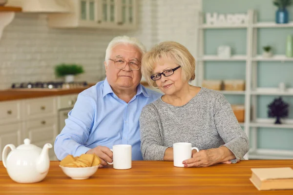 Des grands-parents retraités souriants assis ensemble dans la cuisine avec du thé et des biscuits — Photo