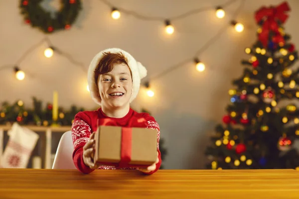 Cute smiling boy sits at a table and holds out a Christmas present in front of him giving it. — Stock Photo, Image