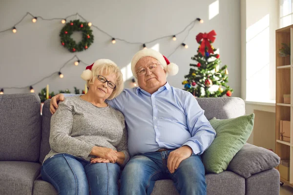 Heureux couple de personnes âgées dans chapeaux de Père Noël regardant la caméra tout en étant assis sur le canapé à la maison — Photo