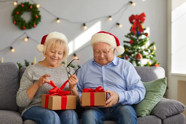 Noel şapkalı mutlu çift koltukta oturuyor ve bayram hediyelerini açıyor. — Stok fotoğraf