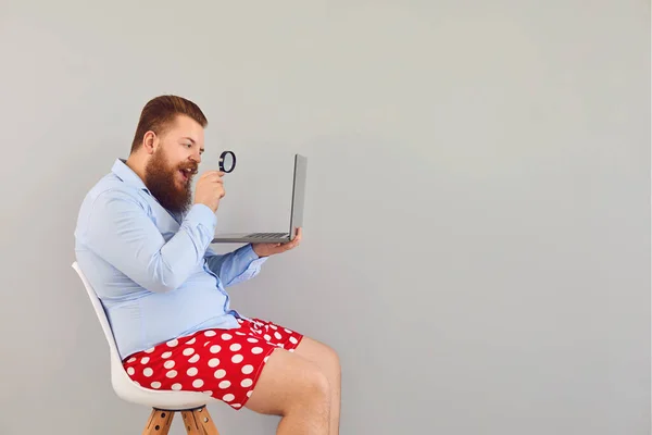 Lustiger Mann in roten Shorts mit Laptop vergrößert die Informationen auf grauem Hintergrund. Konzept der Online-Freelancer, die aus der Ferne arbeiten. — Stockfoto