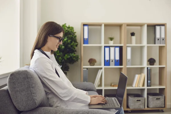 Smiling woman doctor sitting in clinic with laptop on knees and communicating during online consultation — Stock Photo, Image