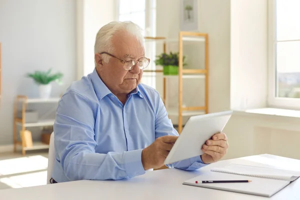 タブレットを使用して自宅でテーブルに座ってメガネを持つシニア男は、インターネット上の情報を読み取ります. — ストック写真