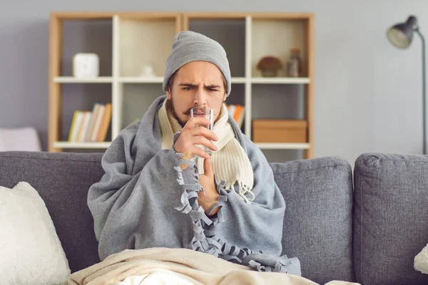 Malato infelice seduto sul divano avvolto nella coperta e che prende la medicina con acqua — Foto Stock