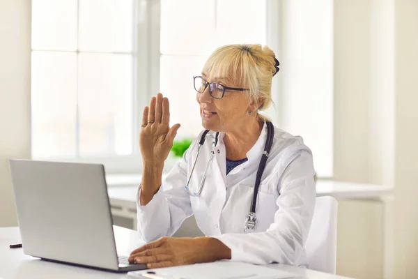 Vista lateral amigable médico senior mujer saluda a la paciente y lleva a cabo una consulta en línea. — Foto de Stock