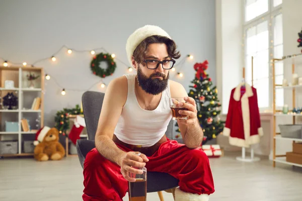 Noel günü evde sandalyede oturan ve viski içen atletli ve Noel Baba şapkalı genç adam. — Stok fotoğraf