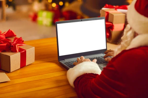 Senior Santa στην εορταστική παραδοσιακή φορεσιά και καπέλο κάθεται δακτυλογράφηση Χριστούγεννα συγχαρητήρια σε απευθείας σύνδεση για laptop — Φωτογραφία Αρχείου