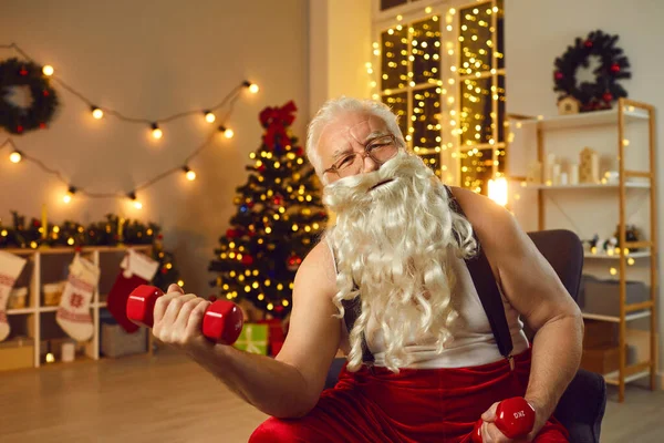 Szczęśliwy Święty Mikołaj siedzi w fotelu w salonie i ćwiczy z hantlami — Zdjęcie stockowe