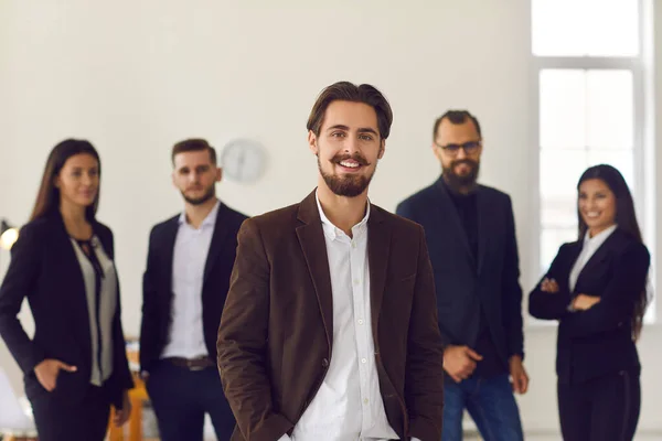 Usmívající se podnikatel nebo majitel společnosti při pohledu do kamery nad pozitivními kolegy na pozadí — Stock fotografie