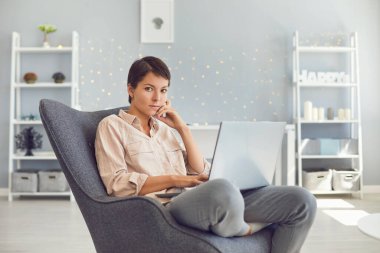 Dizüstü bilgisayarla çalışırken kameraya bakan ciddi bir kadın rahat koltukta oturuyor.