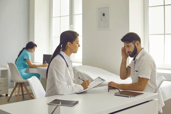 Clínica médica. Mujer doctora escuchando mostrando dolor de cabeza hombre paciente y tomando notas —  Fotos de Stock