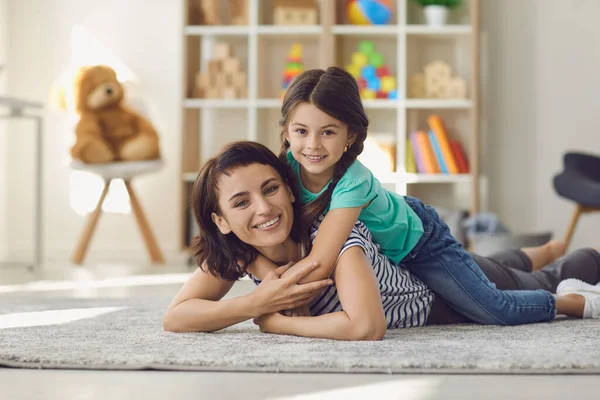 Smiling daughter lying on happy mother back on karpet and looking at camera — Stok Foto